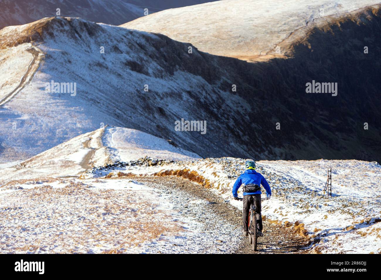 A mountain Biker descending a rocky trail from the summit of Grisedale Pike towards Sand Hill and Hopegill Head in winter in the English Lake District Stock Photo