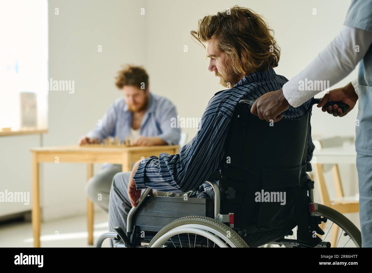 Hands of personnel of mental hospital pushing wheelchair with male patient suffering from