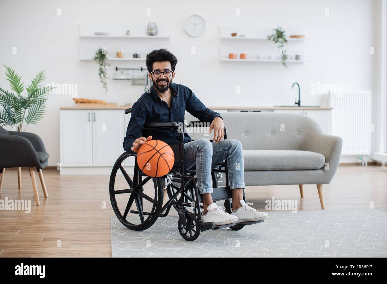 Cheerful bespectacled adult with mobility impairment dribbling ball in  studio apartment on weekend. Happy indian in jeans and dark shirt staying  active with disability while playing game for pleasure Stock Photo -