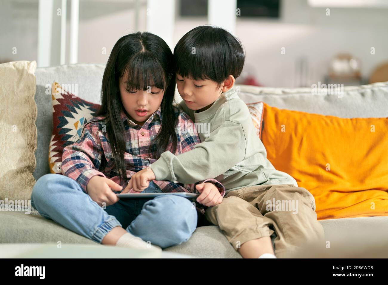 two asian children brother and sister sitting on family couch at home playing computer game using digital tablet Stock Photo