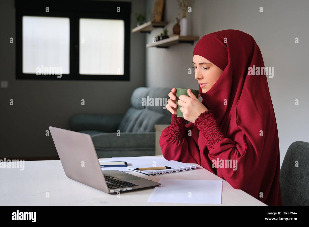 Muslim young woman in hijab drinking coffee while working in a laptop ...