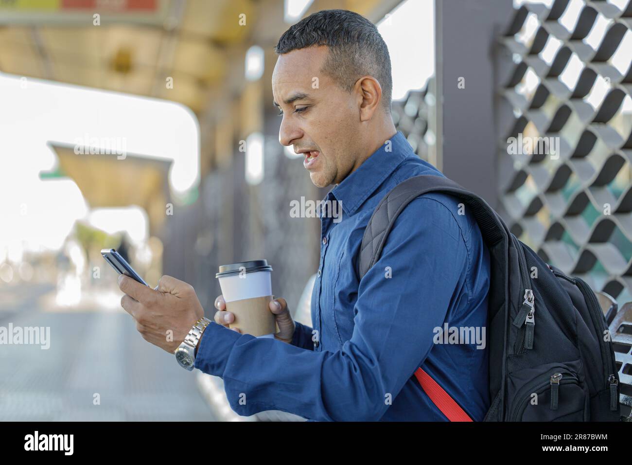 Young latin man looking at his mobile phone with a surprised face a paper cup with coffee in his hand. Stock Photo