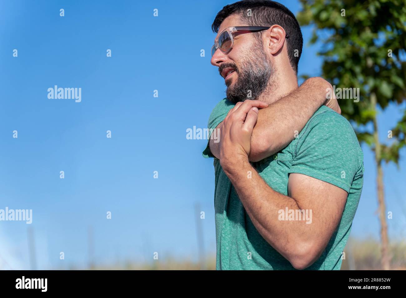 Profile view of an attractive bearded man with sunglasses stretching his shoulders in a field with blue sky in the background Stock Photo