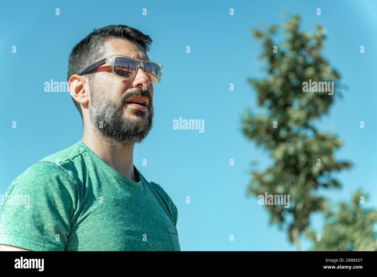 Attractive young man with beard and sunglasses in profile wearing a green t-shirt looking at the sun with a tree in the background Stock Photo