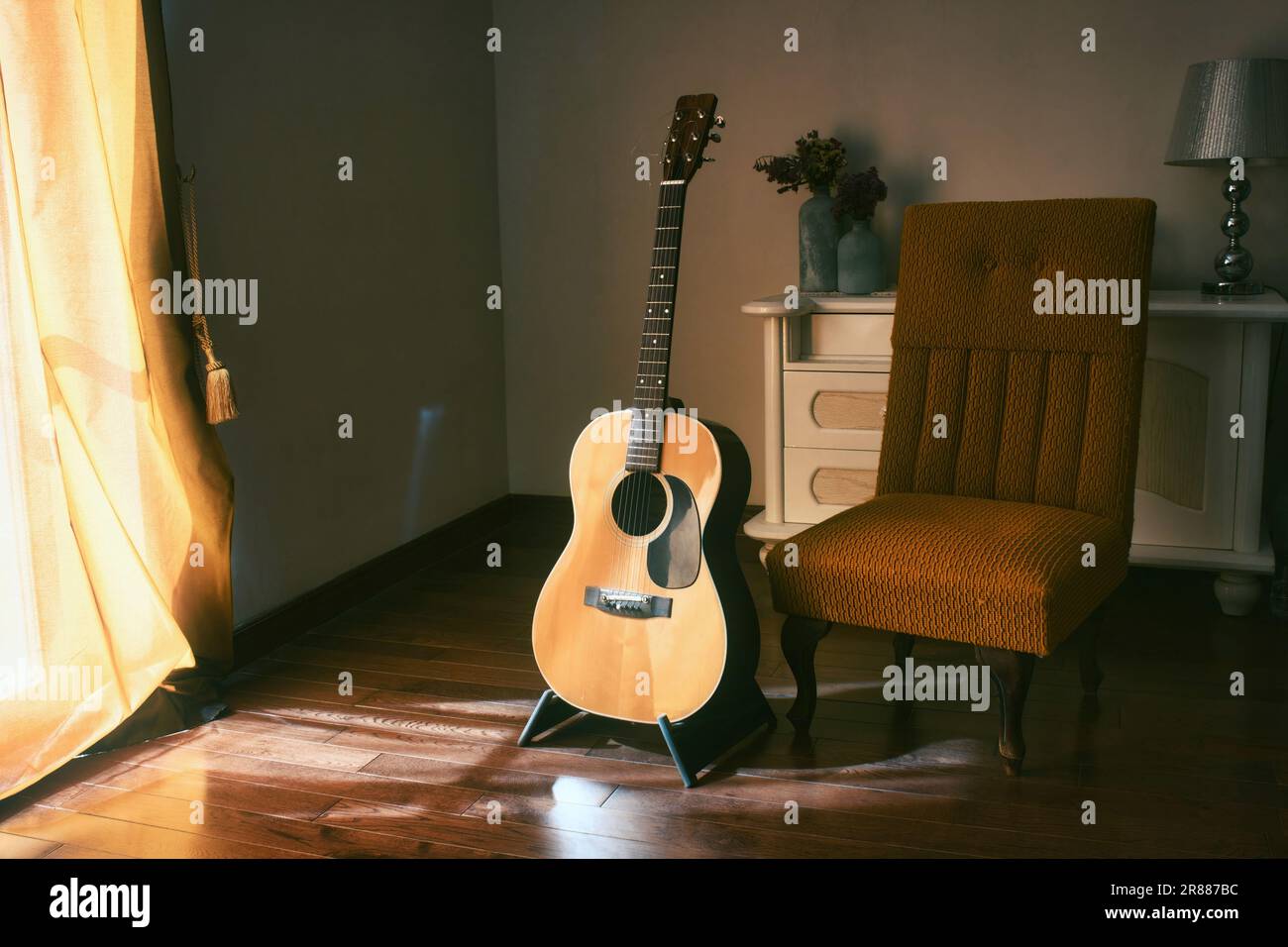 An acoustic Spanish guitar on a stand next to a chair in the moody shadows of a dark room with bright light coming in from behind a curtain Stock Photo