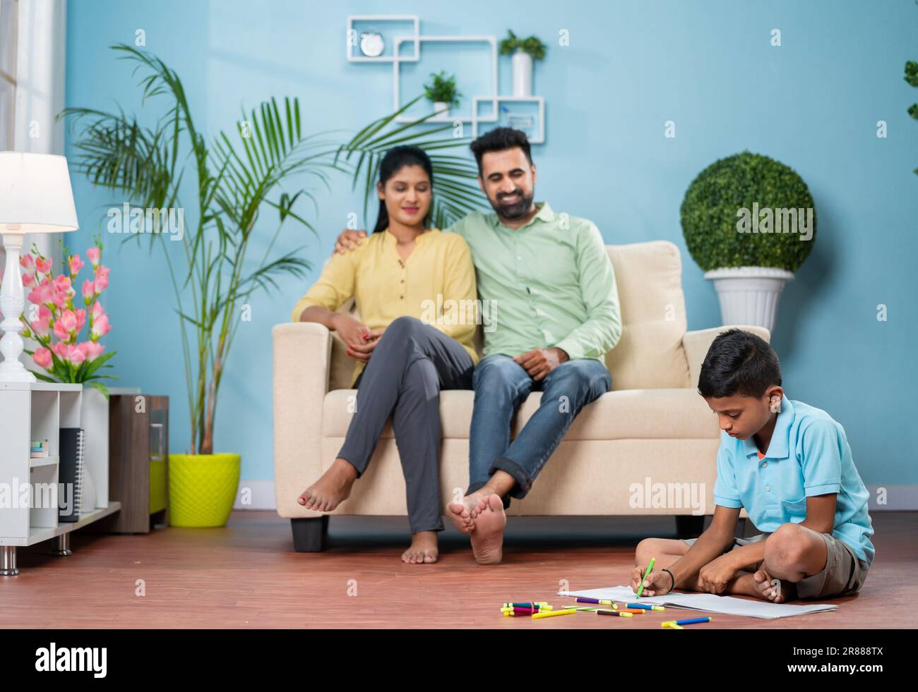 Happy indian kid drawing with pencil sitting on floor in front of couple on sofa at home Stock Photo