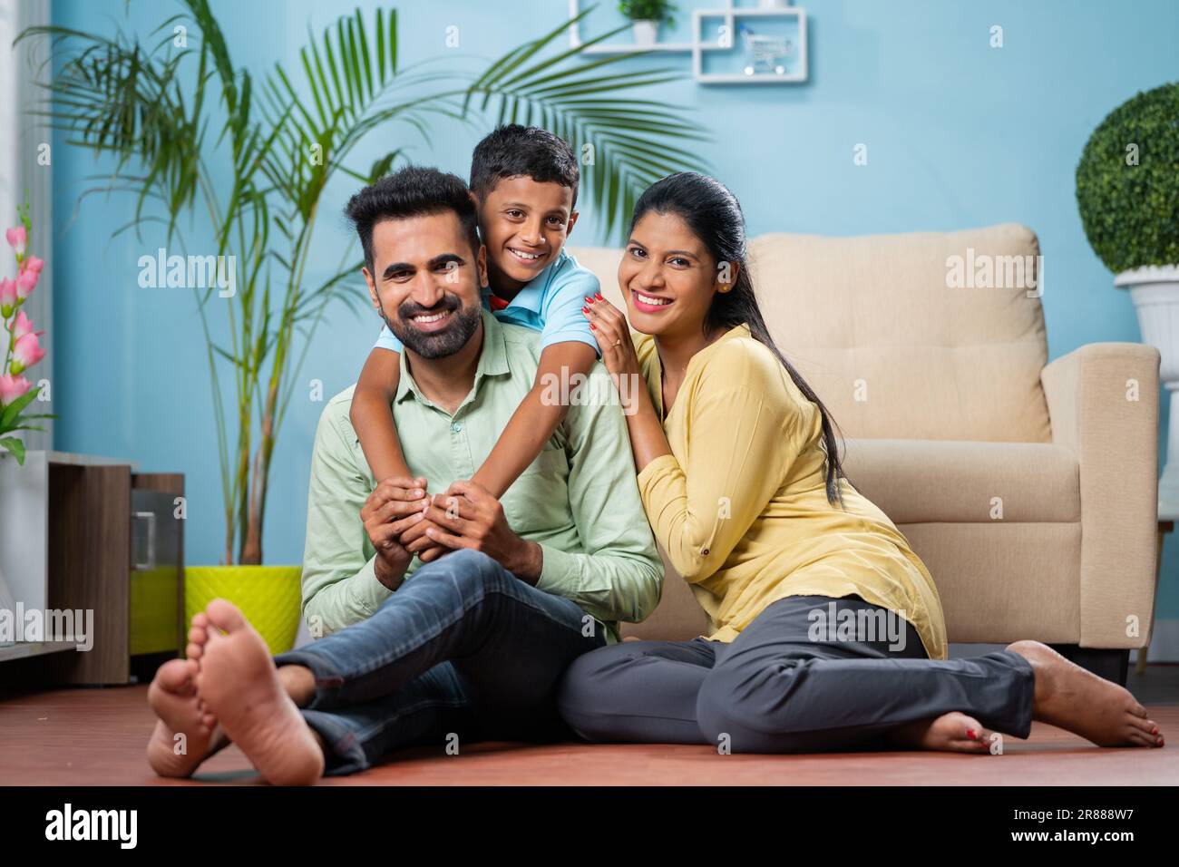 Happy indian parents with son by looking at camera while sitting on floor at home - concept of family bonding, weekend holiday and parenthood. Stock Photo