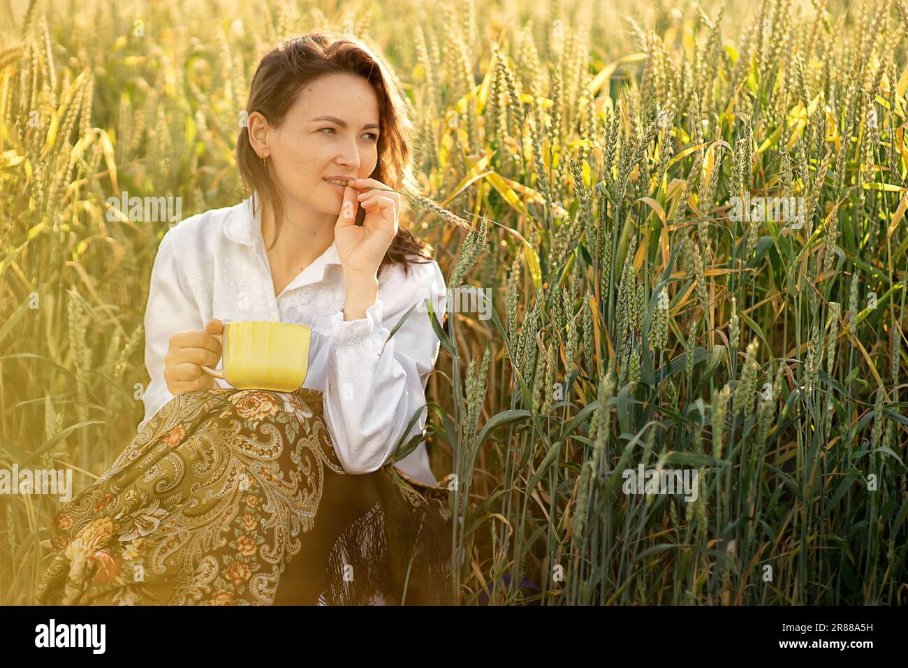 a young white woman went out at dawn to a wheat field to drink a cup of tea, she is sitting on the edge of the field, she has a spikelet in her mouth, Stock Photo