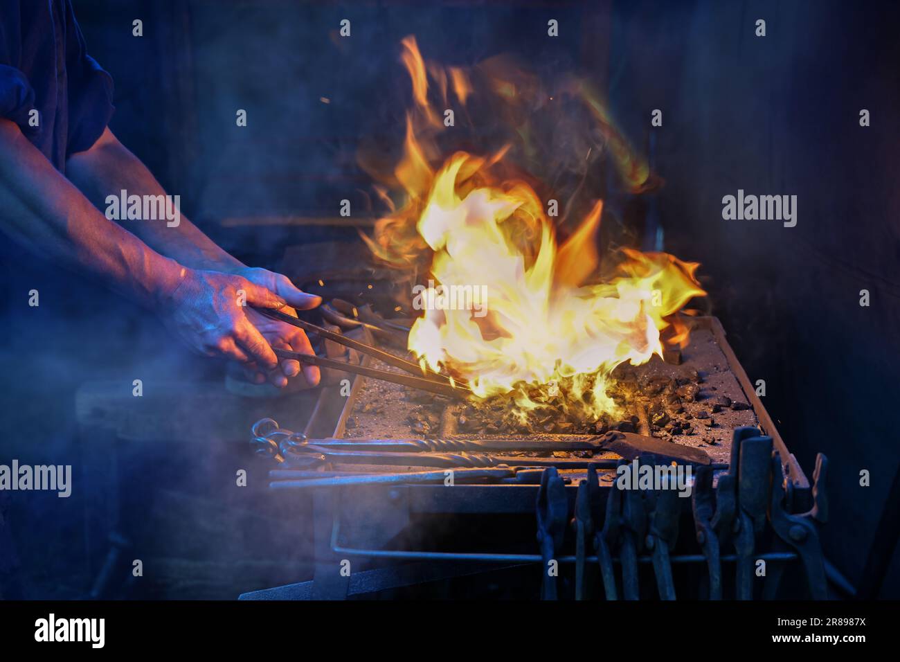 Hands of a blacksmith holding tongs with a workpiece in the blazing fire on the forge to get the iron glowing, smoke in the dark background, copy spac Stock Photo