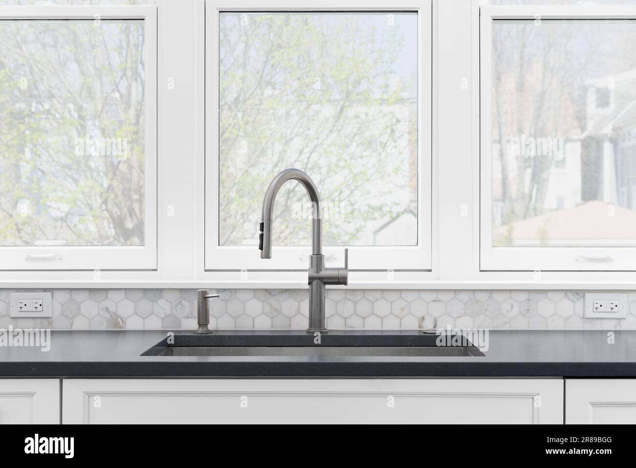 A kitchen sink detail with white cabinets, grey countertop, a marble hexagon backsplash, and large windows. Stock Photo