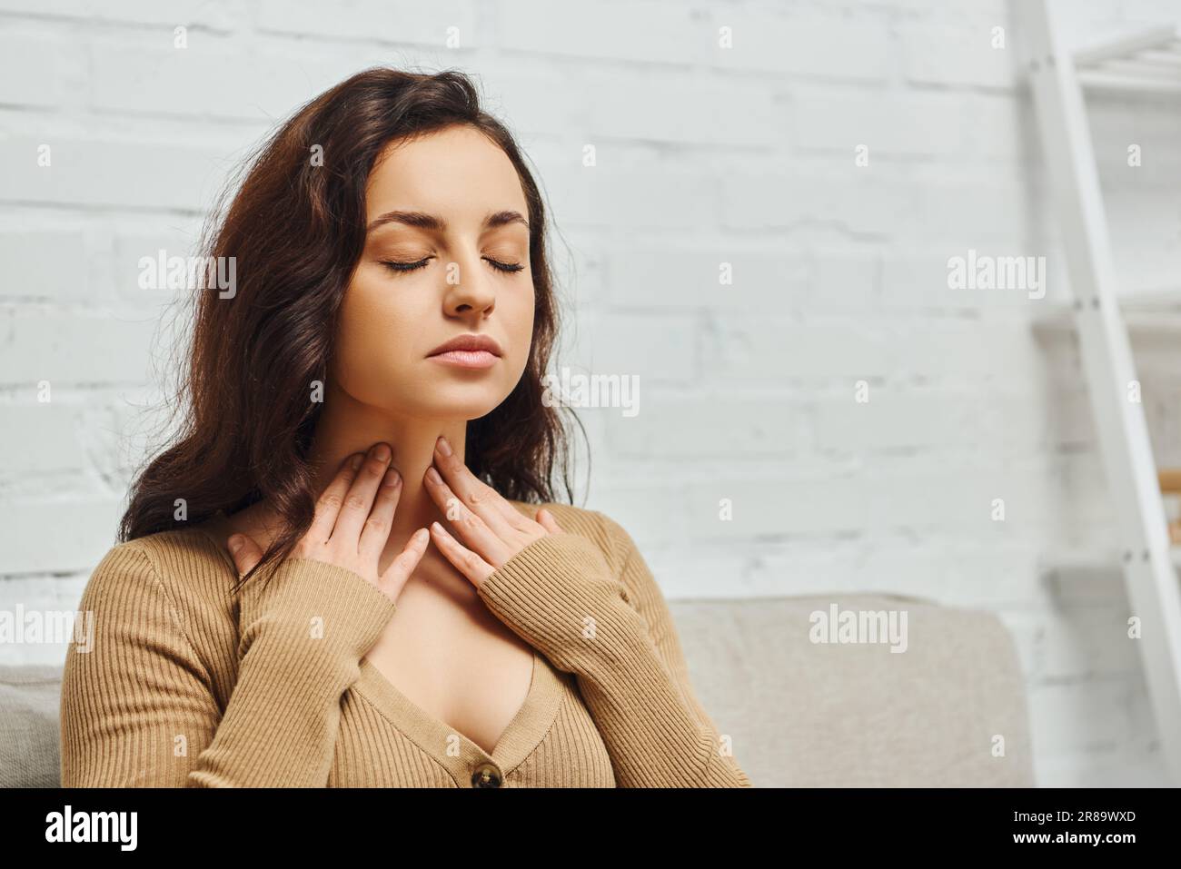 Young brunette woman in jumper checking thyroid gland on neck while sitting with closed eyes on couch in modern living room at home, focus on self-car Stock Photo