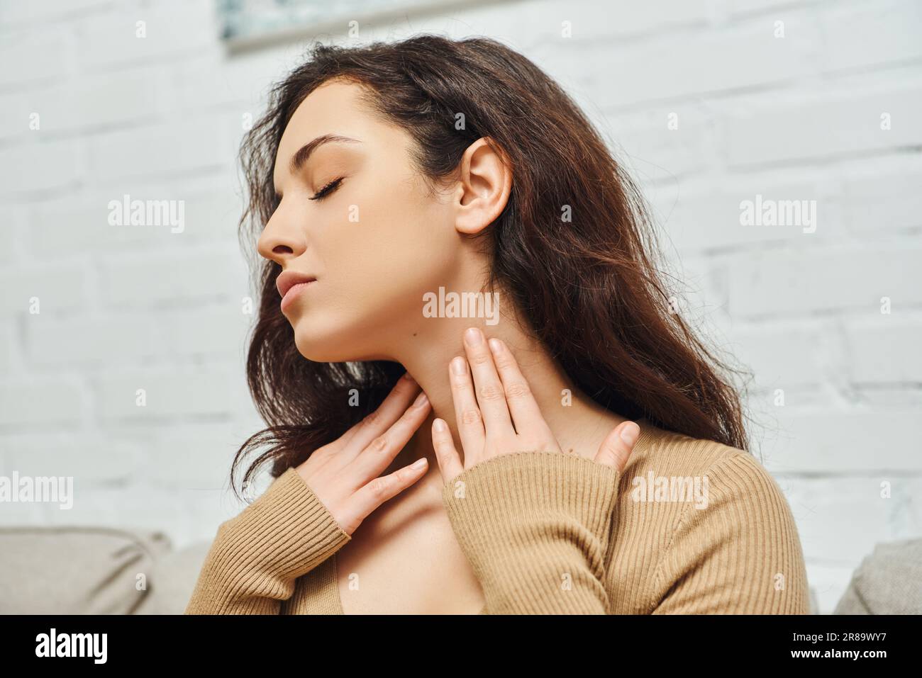 Young brunette woman in casual clothes touching neck while checking thyroid gland and sitting on couch in living room at home, focus on self-care and Stock Photo