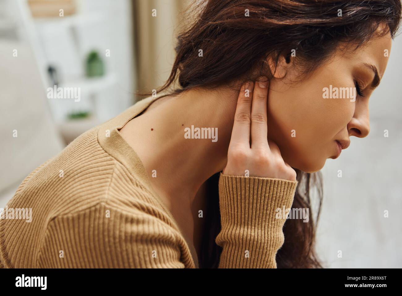 Side view of young brunette woman suffering from pain while doing lymphatic self-massage in blurred house, self-care ritual and holistic healing conce Stock Photo