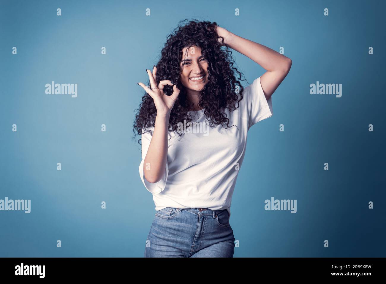 Shot of happy confident curly woman with toothy smile, wears casual basic solid white t-shirt, expresses good emotions, enjoys nice day, doing ok symb Stock Photo