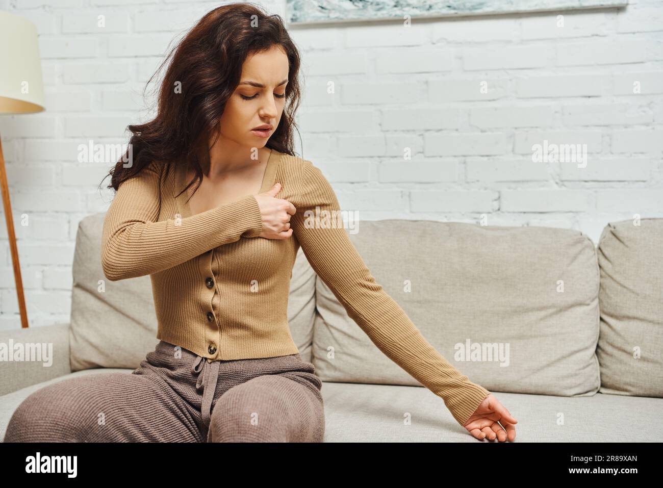 Young brunette woman in brown jumper suffering from pain while massaging lymphatic nodes on armpit and sitting on couch at home, self-care ritual and Stock Photo