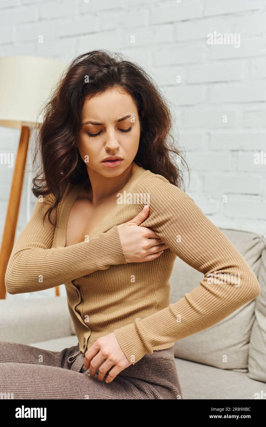 Portrait of young brunette woman in in casual brown jumper massaging lymphatic nodes on armpit during mindful self-care on couch at home, enhancing se Stock Photo