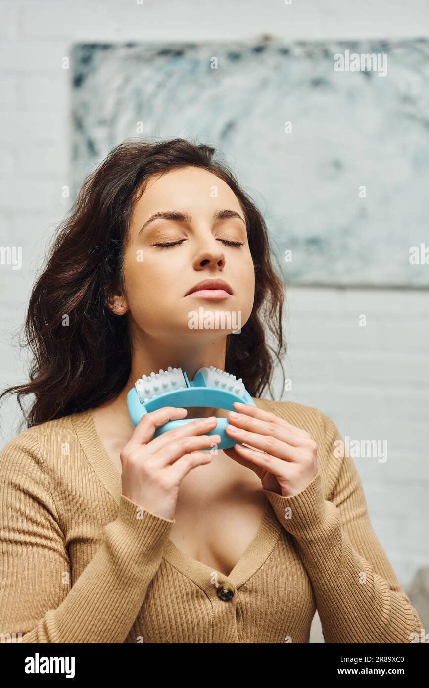 Portrait of young woman with closed eyes in casual brown jumper massaging neck and lymphatic nodes with handled massager at home, enhancing self-aware Stock Photo