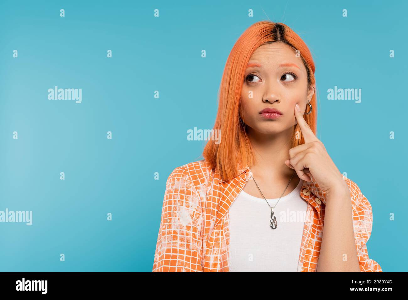 asian woman thinking and looking away, young fashion model touching cheek with finger on blue background, pensive, orange shirt, generation z, vibrant Stock Photo