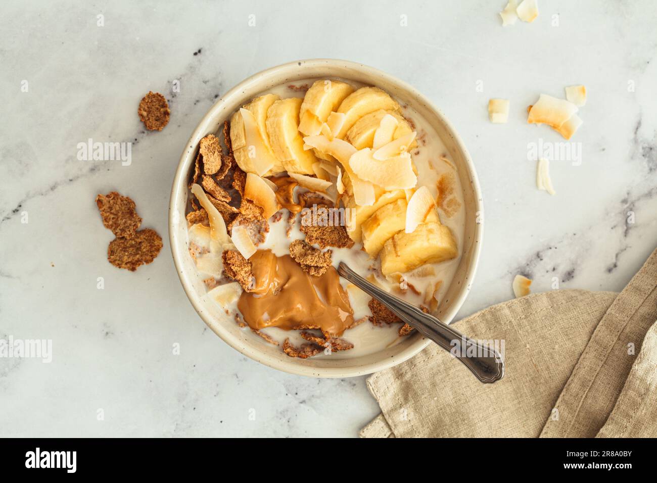 Whole grain flakes with banana, coconut chips and peanut butter in a bowl, white background, top view. Healthy breakfast concept. Stock Photo