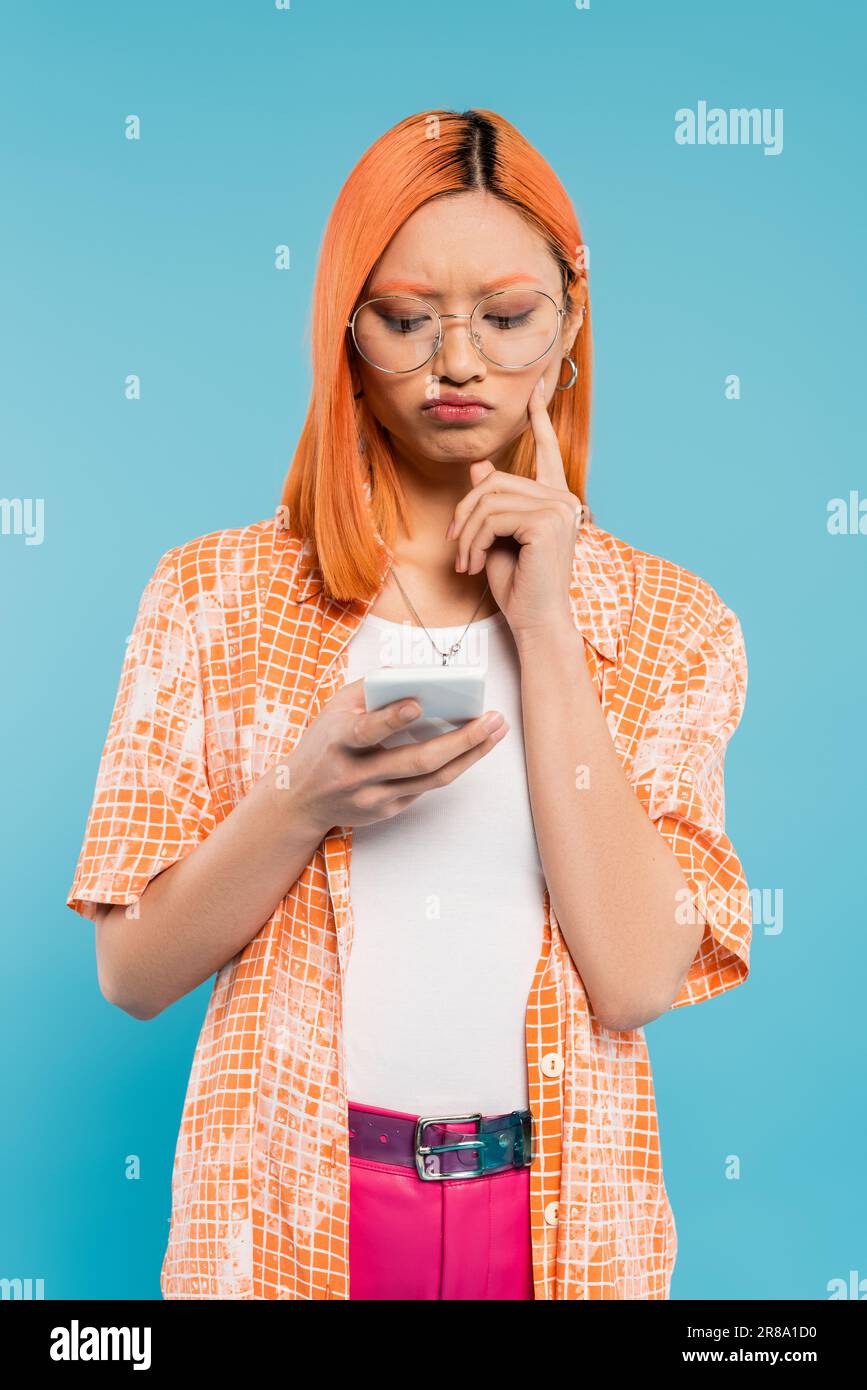 negative emotion, bad mood, displeased asian woman touching cheek while looking at smartphone on blue background, trendy eyeglasses, red colored hair, Stock Photo