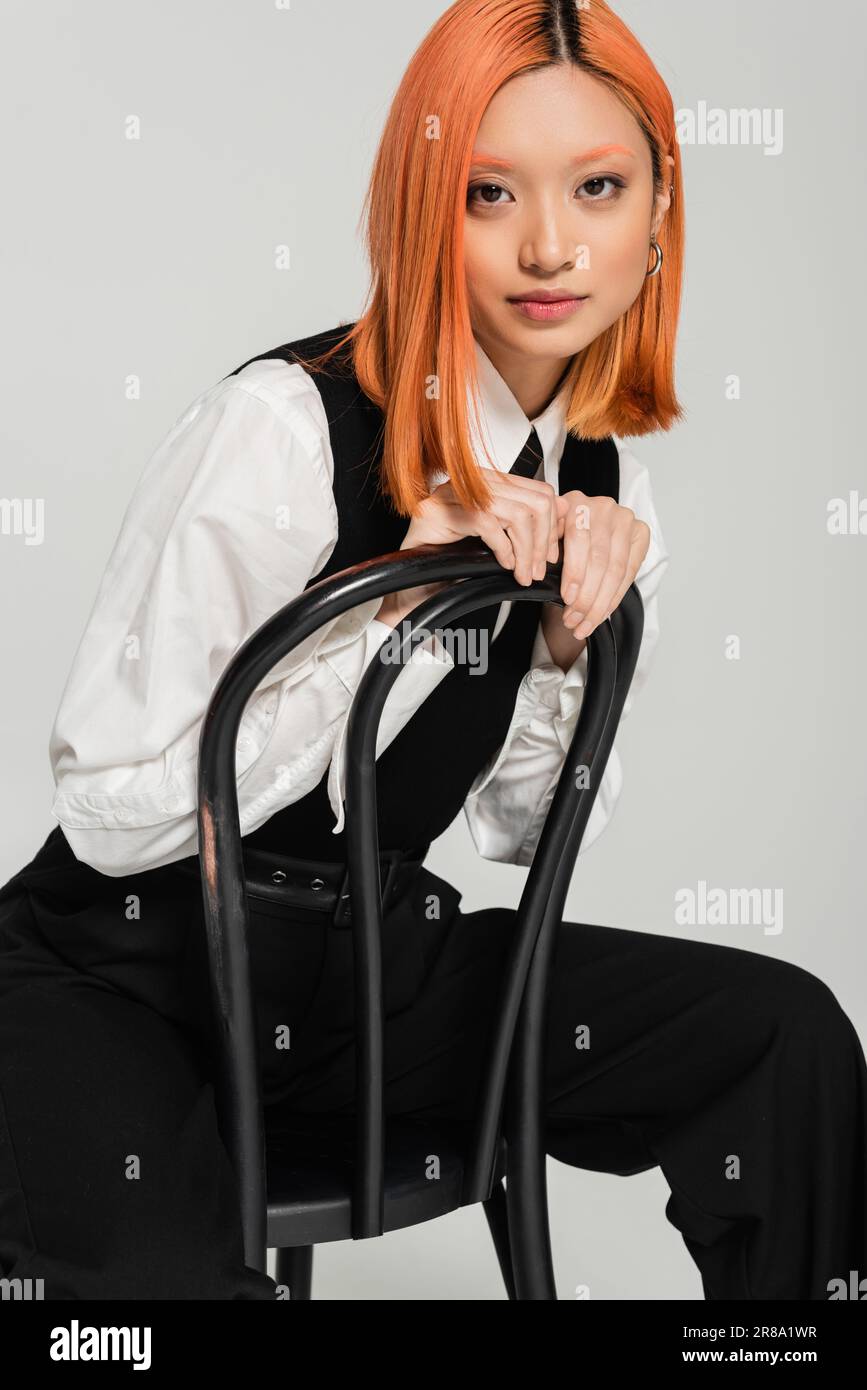 charismatic and fashionable asian woman with colored red hair, white shirt, black vest and pants posing on chair and looking at camera on grey backgro Stock Photo