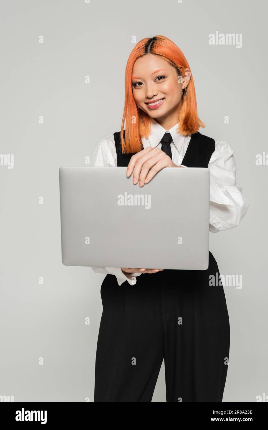 happiness, freelance lifestyle, attractive asian woman in black and white clothing holding laptop and smiling at camera on grey background, colored re Stock Photo