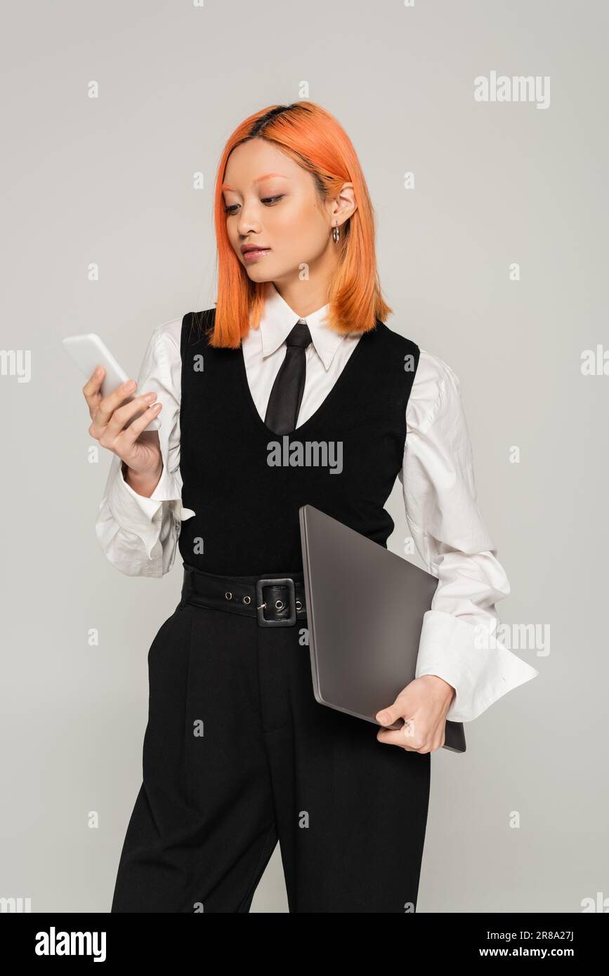 freelance lifestyle, business casual fashion, red haired asian woman in white shirt, black tie and vest standing with laptop and looking at mobile pho Stock Photo