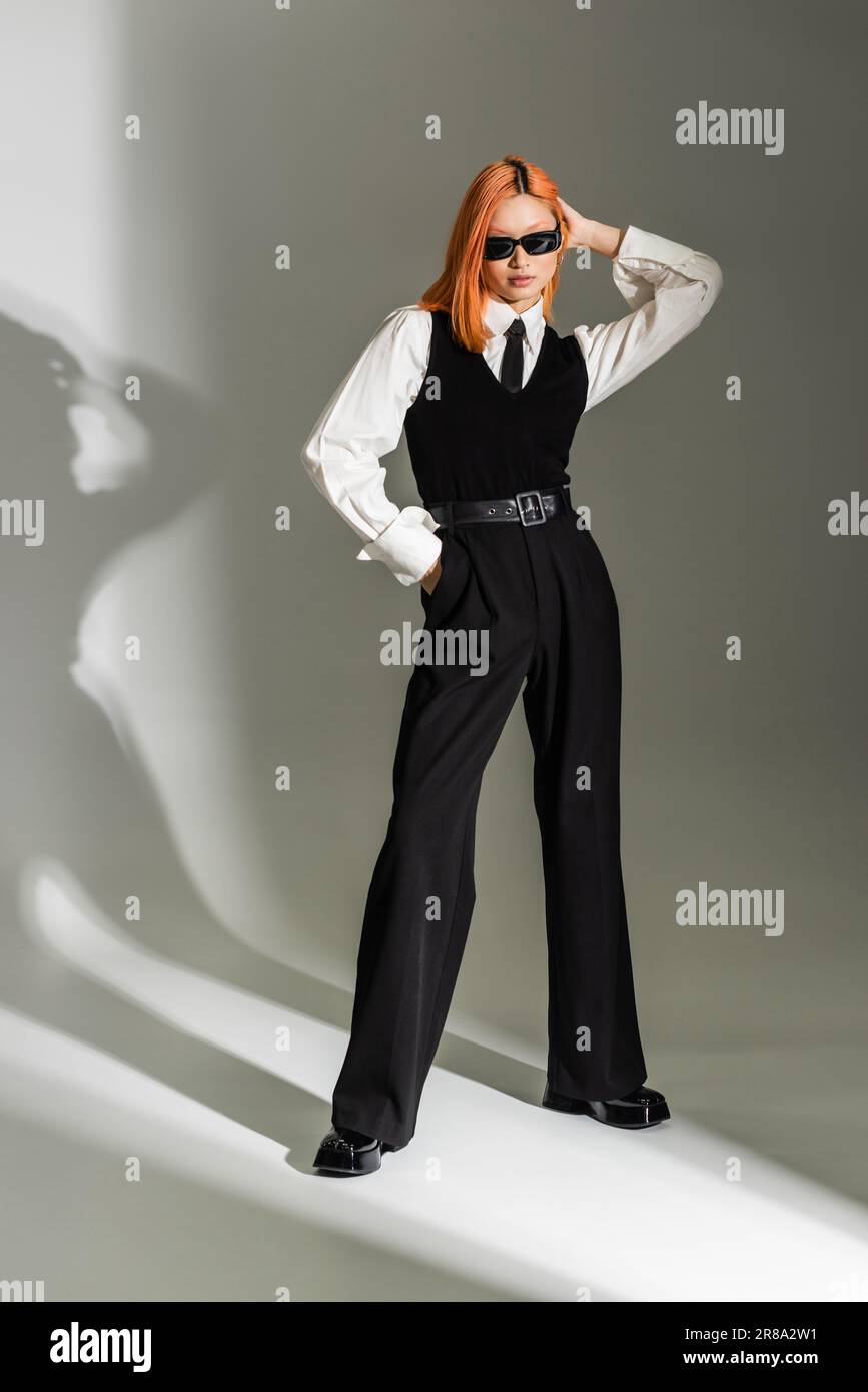 business fashion, trendy asian woman with colored red hair posing with hand in pocket on grey shaded background, dark sunglasses, white shirt, black t Stock Photo