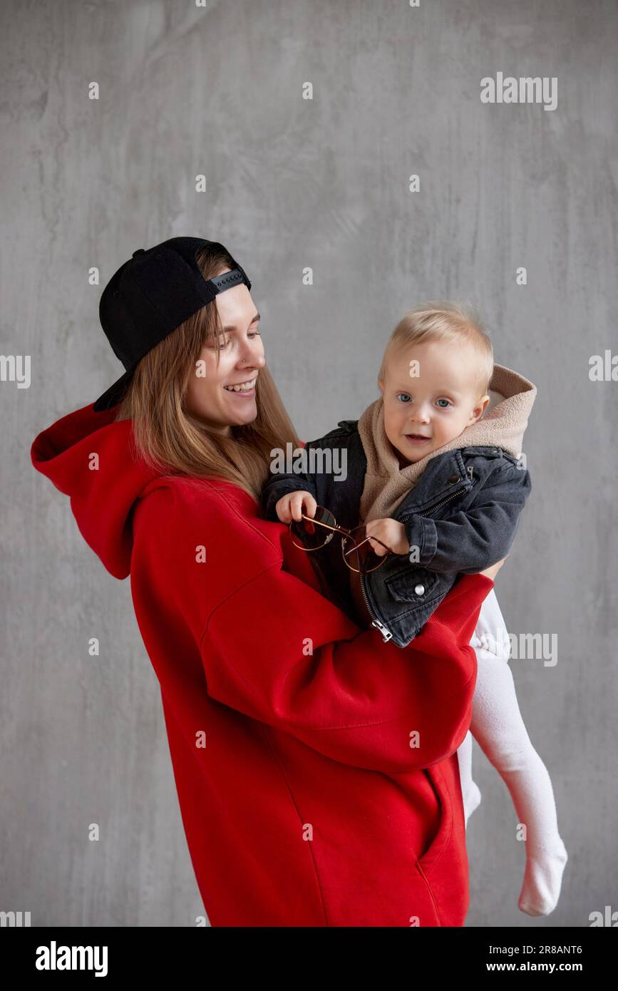 Young cute long haired mom in red hoodie and black baseball cap is holding and hugging her cute baby in beige hoodie and funny motorcycle jacket. Stud Stock Photo Alamy