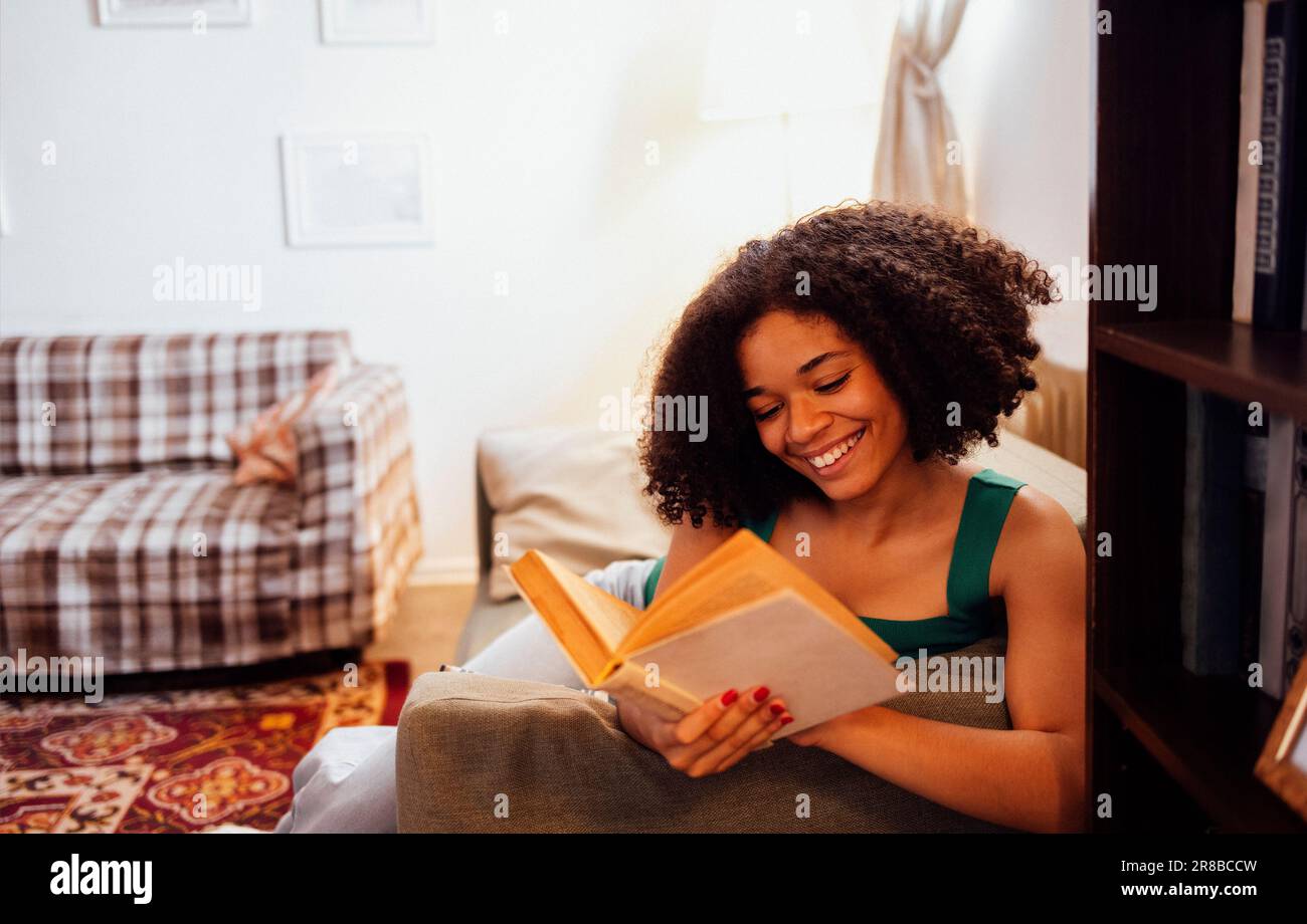Smiling girl of mixed race holds and reads a book on the sofa by