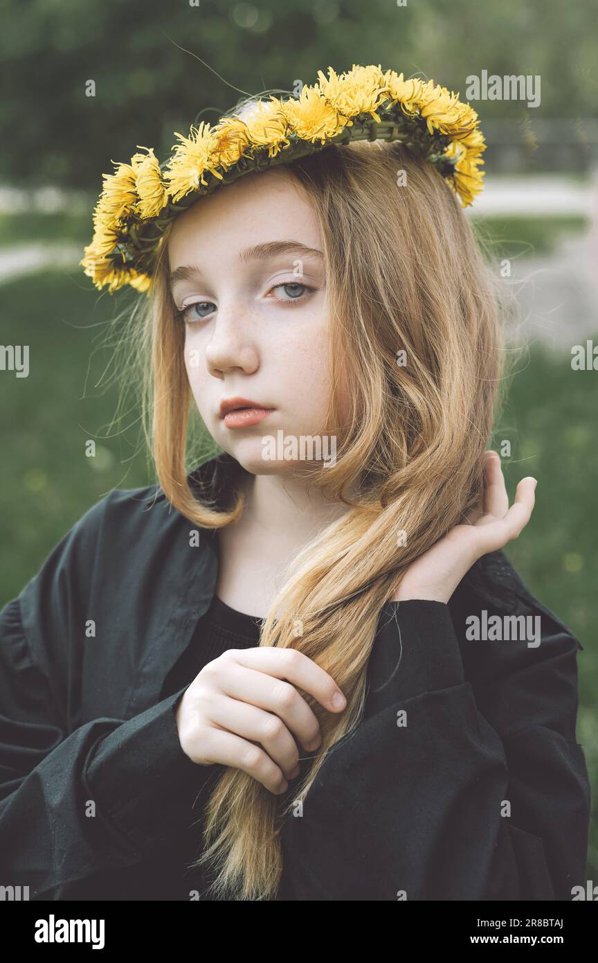 A serious, mysterious girl in a wreath of dandelions. braids a long braid of blond hair. Stock Photo