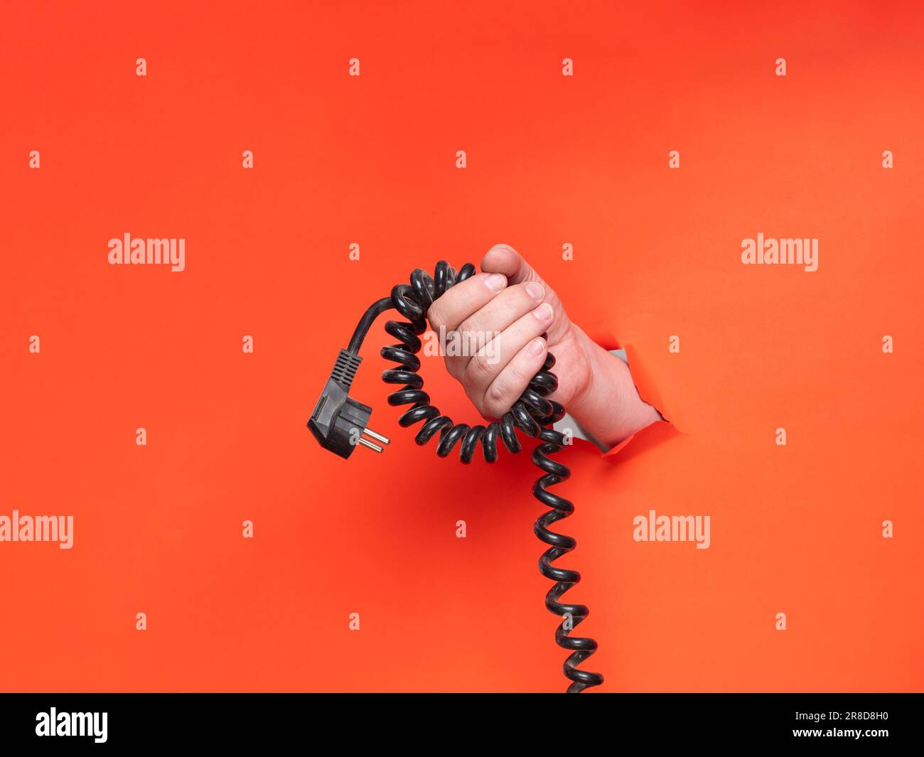 A man's hand holds a black spiral cable through torn orange paper. Concept, hand through torn orange paper. Stock Photo