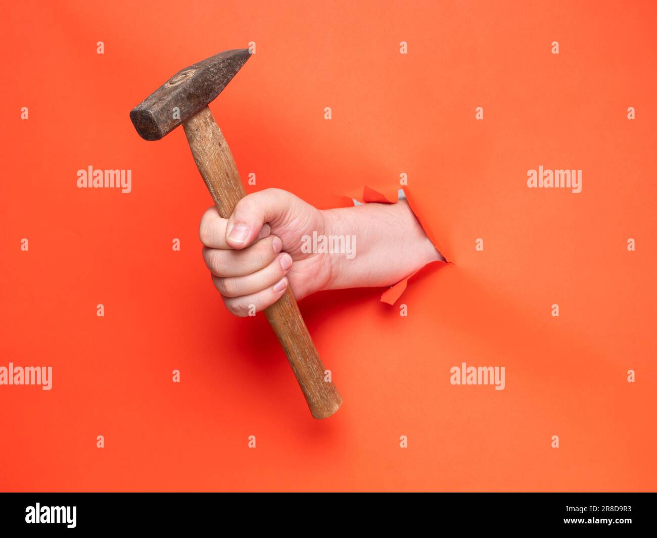 A man's hand holds a hammer through torn orange paper. Concept, hand through torn orange paper. Stock Photo