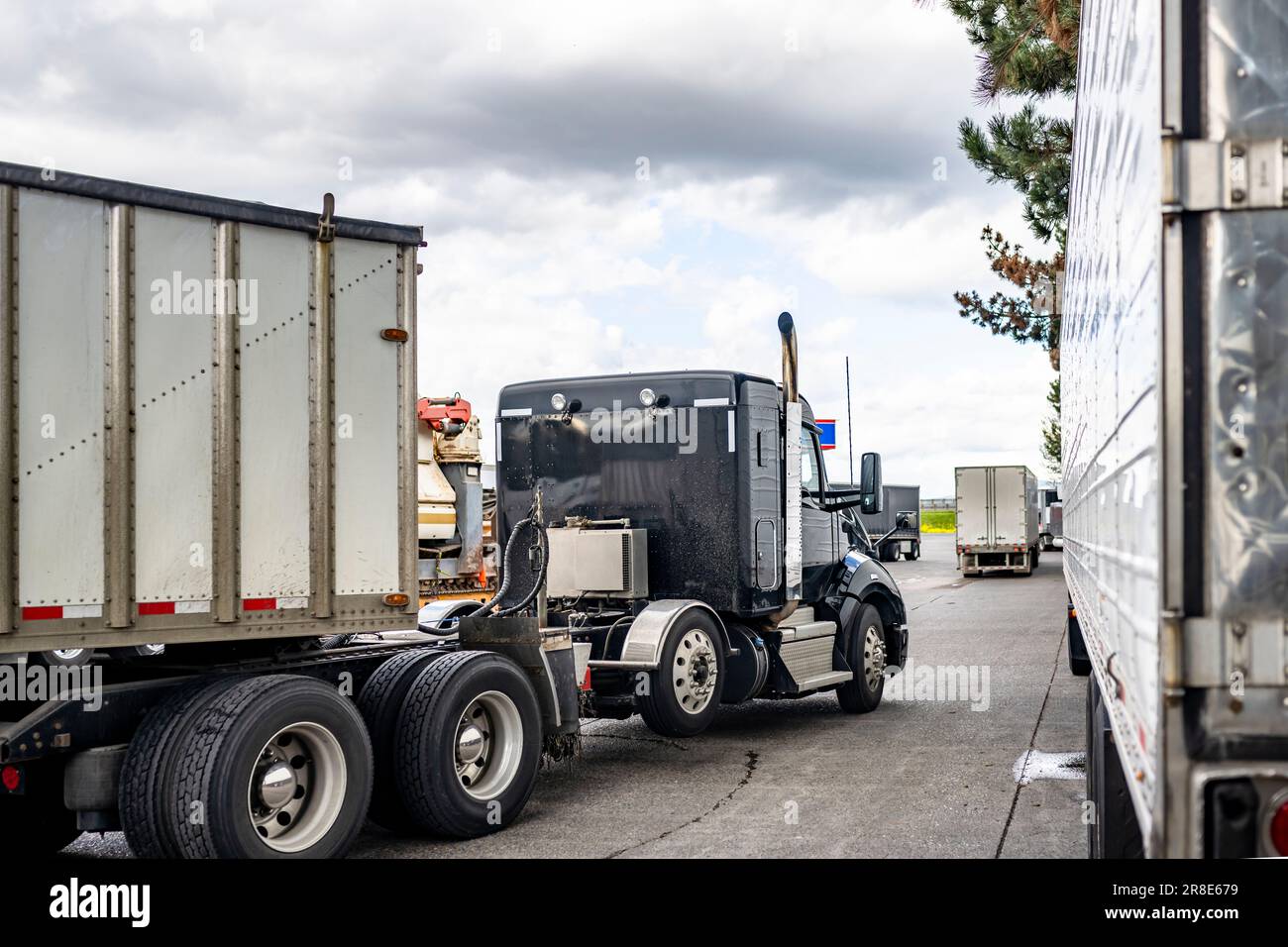 Industrial powerful black big rig classic American bonnet day cab semi truck tractor with bulk semi trailer moving to truck stop parking lot exit with Stock Photo