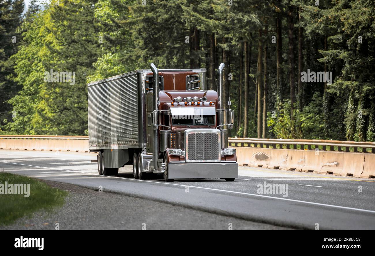 Brown industrial big rig classic American idol semi truck tractor transporting commercial cargo in reefer shiny semi trailer moving on the highway roa Stock Photo