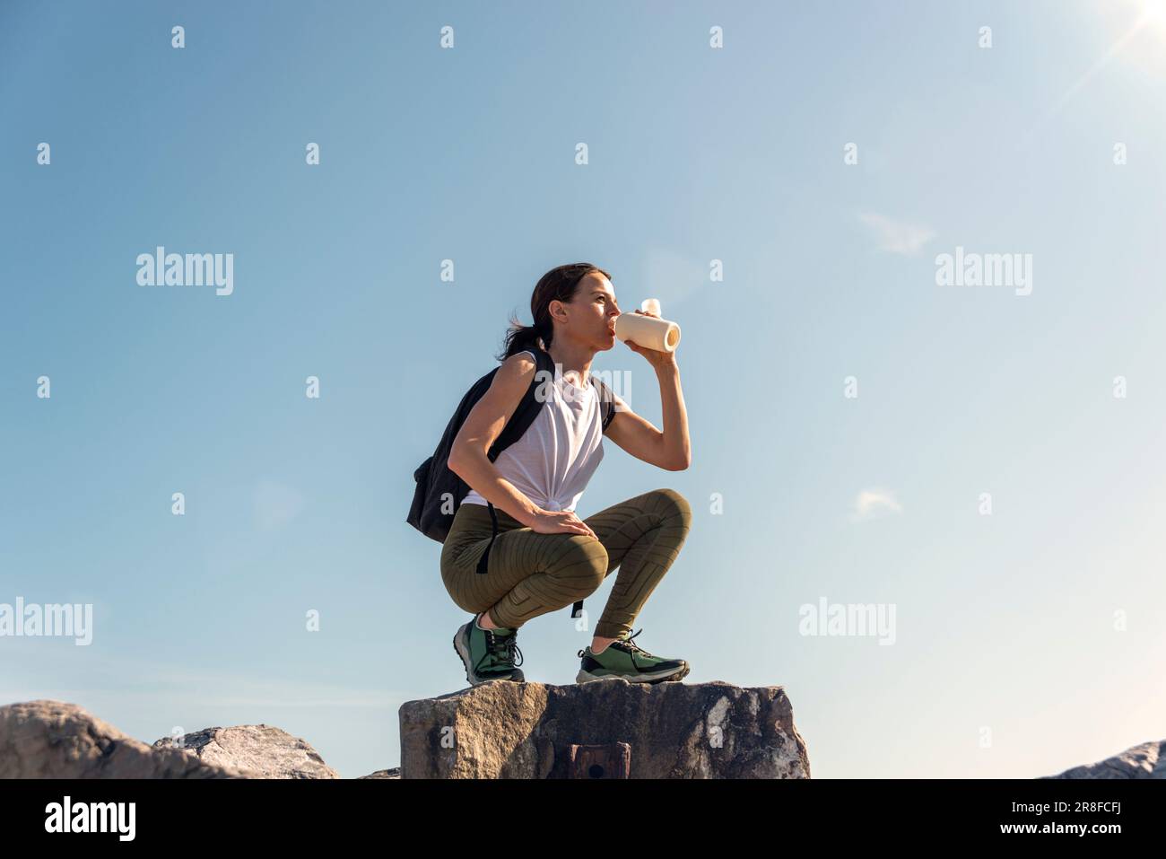 Female walker resting and drinking water on top of rocks with blue sky background. Stock Photo