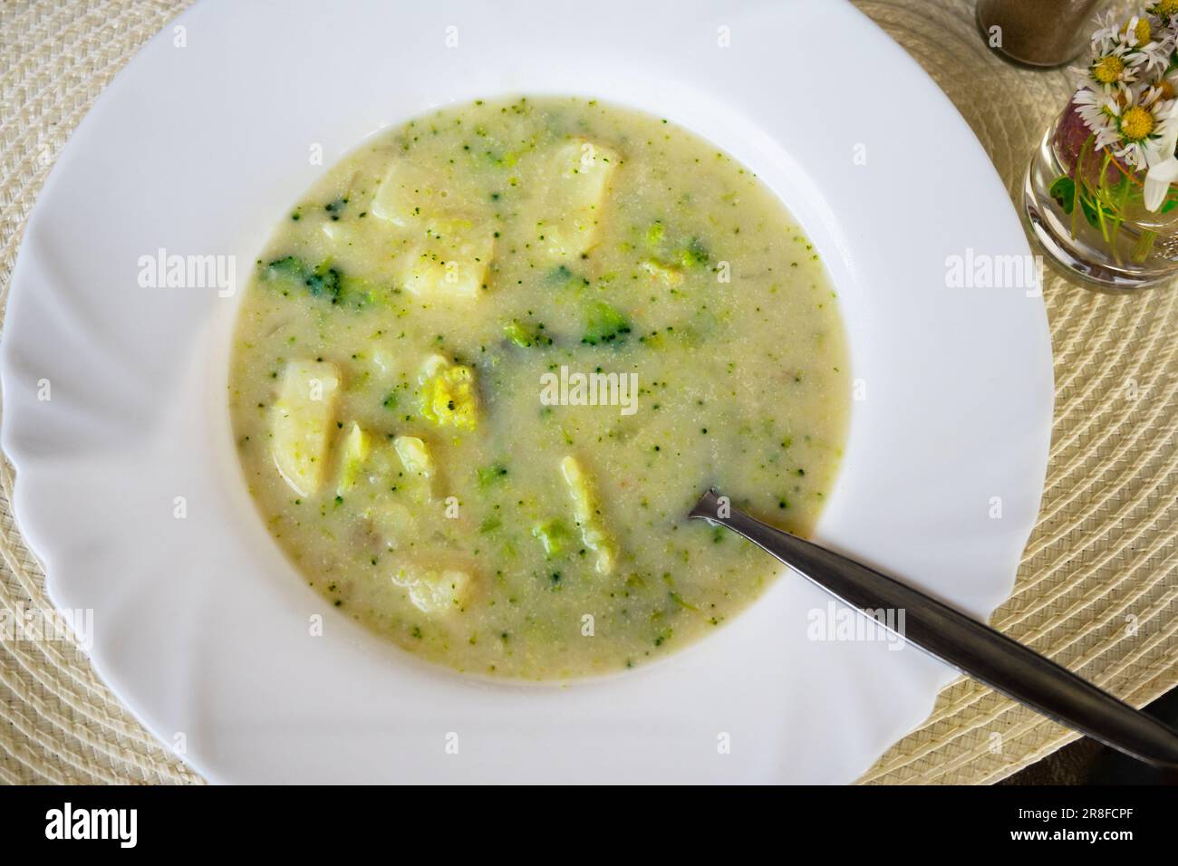 Broccoli soup with sliced potato, spoon in white plate, flowers, pad, closeup. Stock Photo