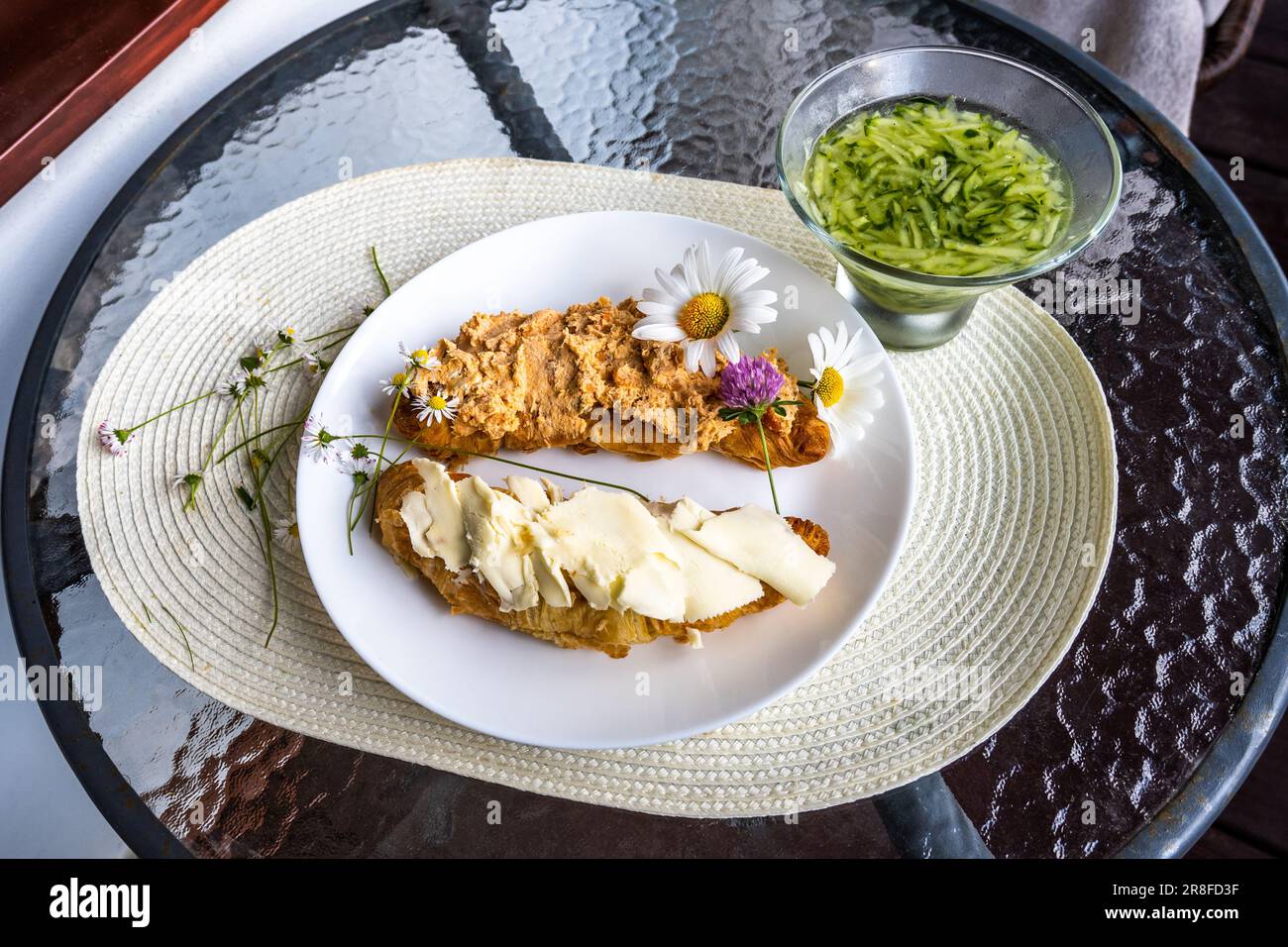 Two piece of croissant with butter and tuna spread on white plate, flowers sprinkled on pad, cucumber salad on circle glass table. Healthy breakfast o Stock Photo
