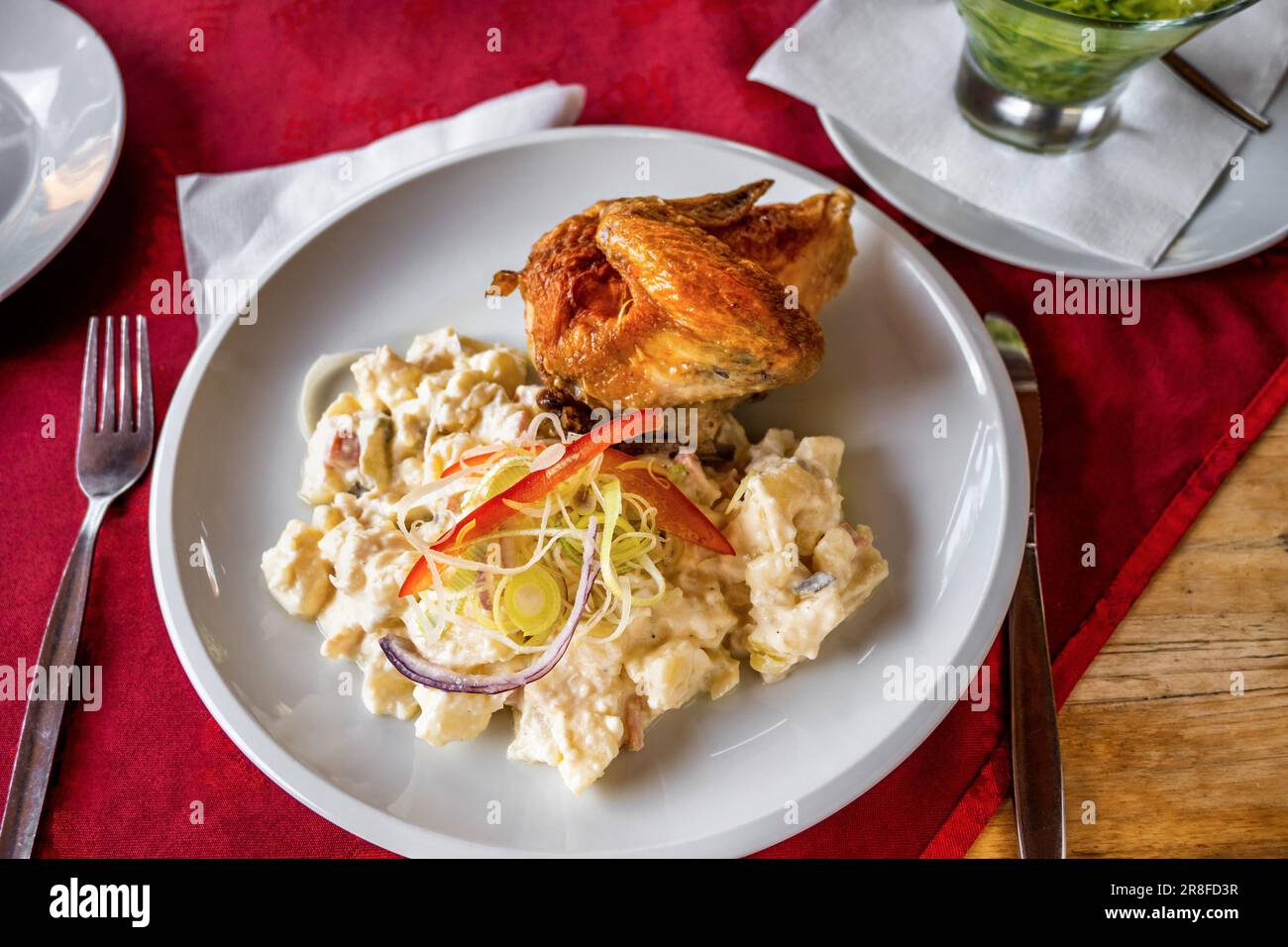 Cold potato salad with mayonnaise and onion, baked chicken breast on white plate, cutlery, part of glass bowl with cucumber salad on red tablecloth, c Stock Photo