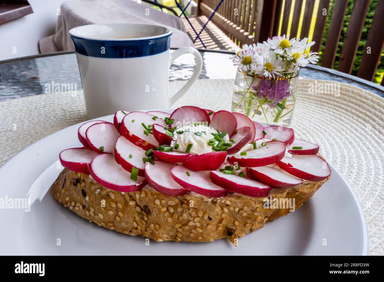 Bread garnished with radish on white plate, cup of coffee, flowers on table, outdoor. Breakfast or snack. Stock Photo