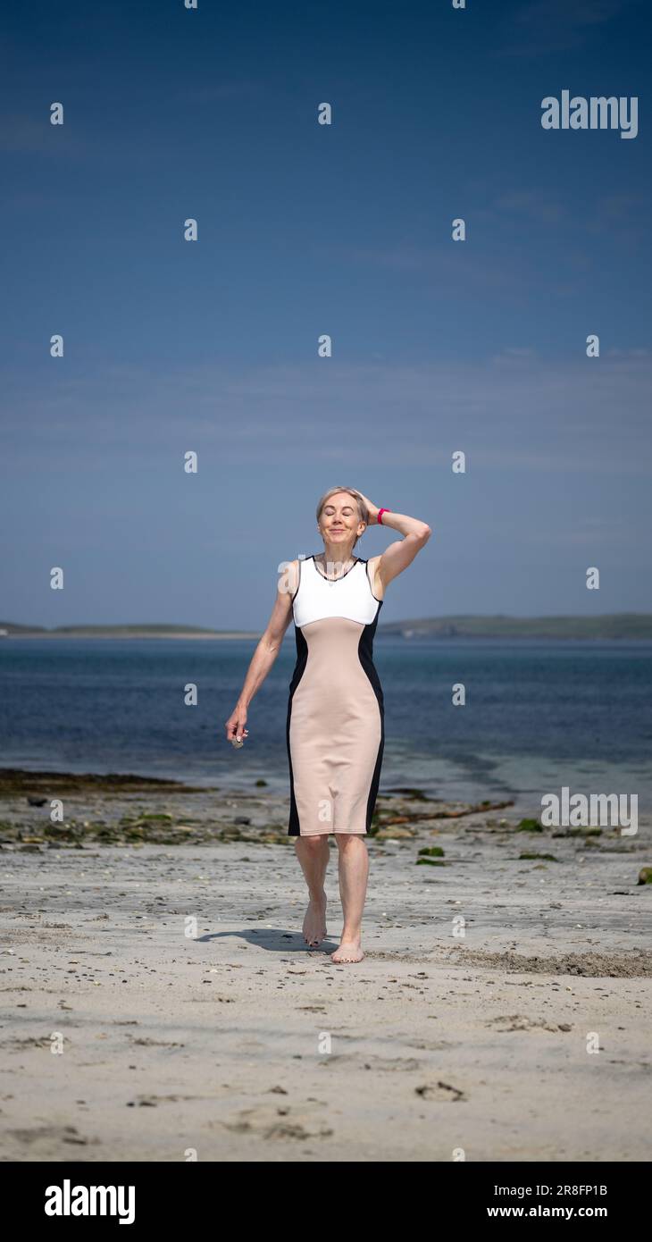 Woman in a smart dress walking barefoot along a sandy beach in the Orkney Isles, Scotland, UK Stock Photo