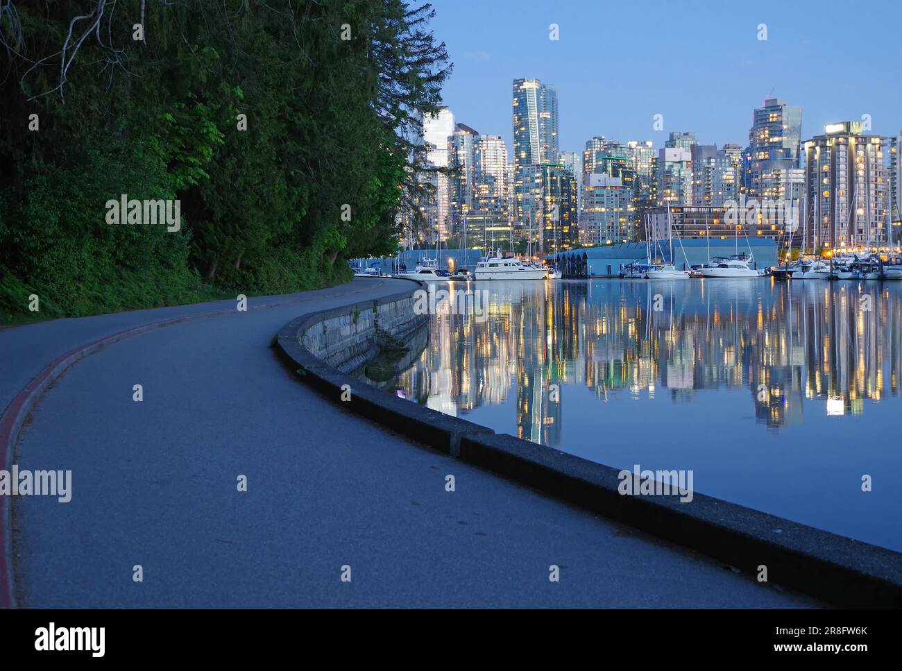 Illuminated skyscrapers and pleasure boats reflected in the calm water, parts of the Sea Wall with bike path in Stanley Park, dusk, Vancouver Stock Photo