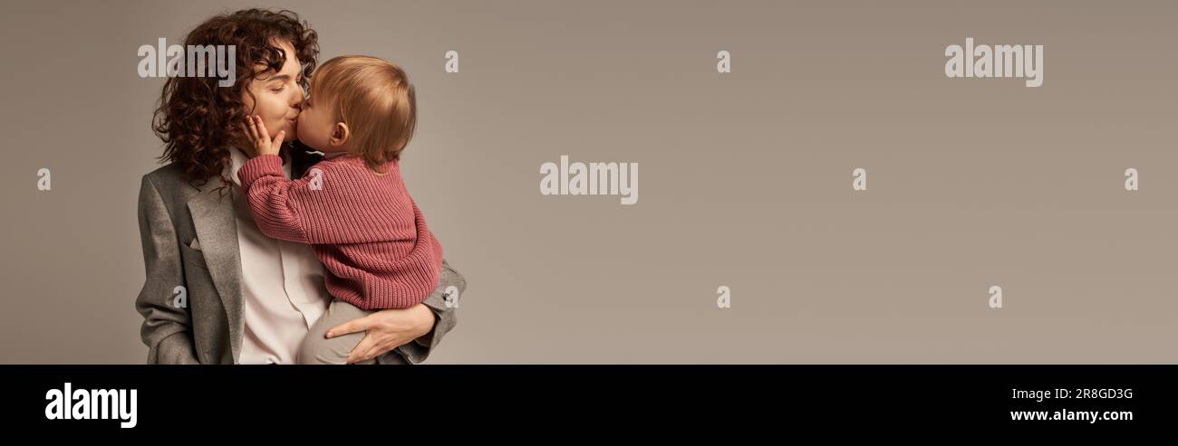 work life balance concept, curly woman in suit kissing daughter on grey ...