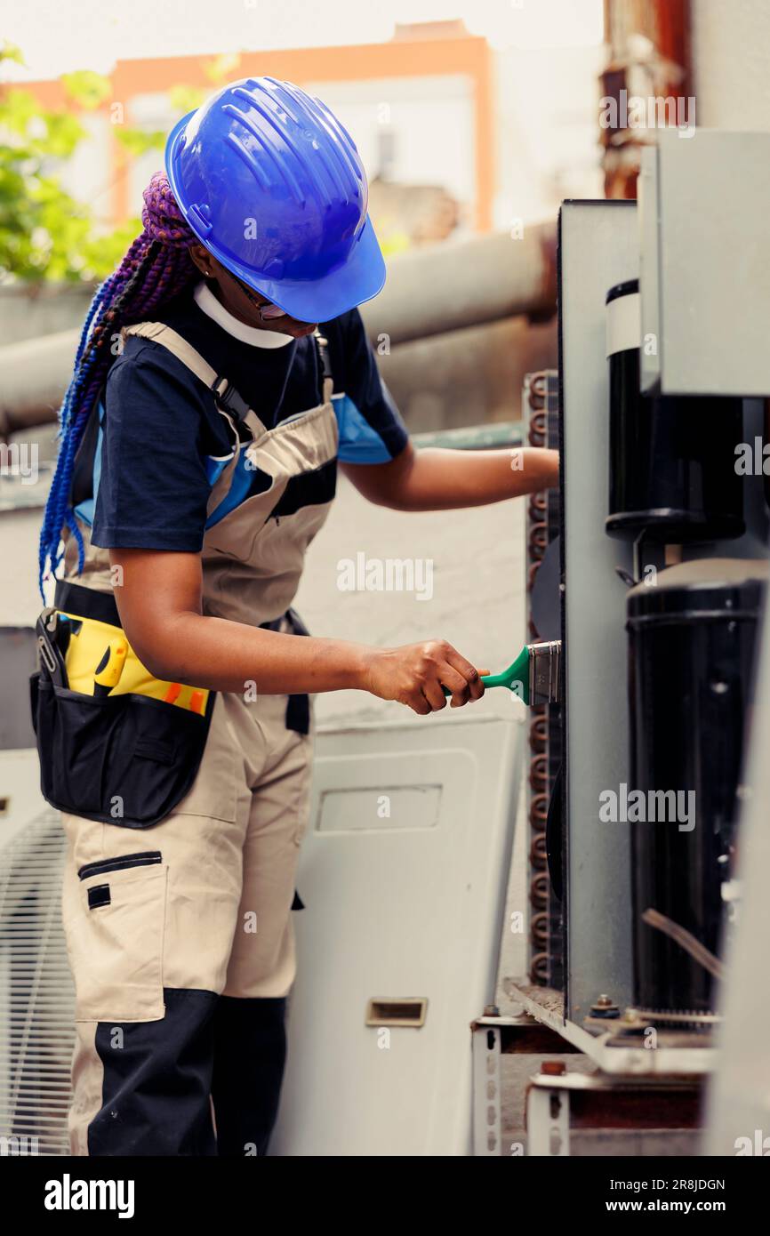African american expert commissioned for annual air conditioner routine cleaning and maintenance, using soft dusting brush to remove built up layer of dirt and dust from compressor coils Stock Photo