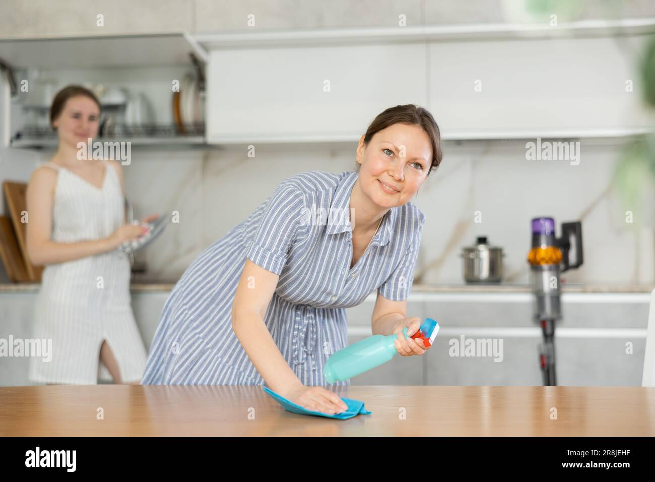 Pleased mother dusting table while daughter washes dishes in kitchen