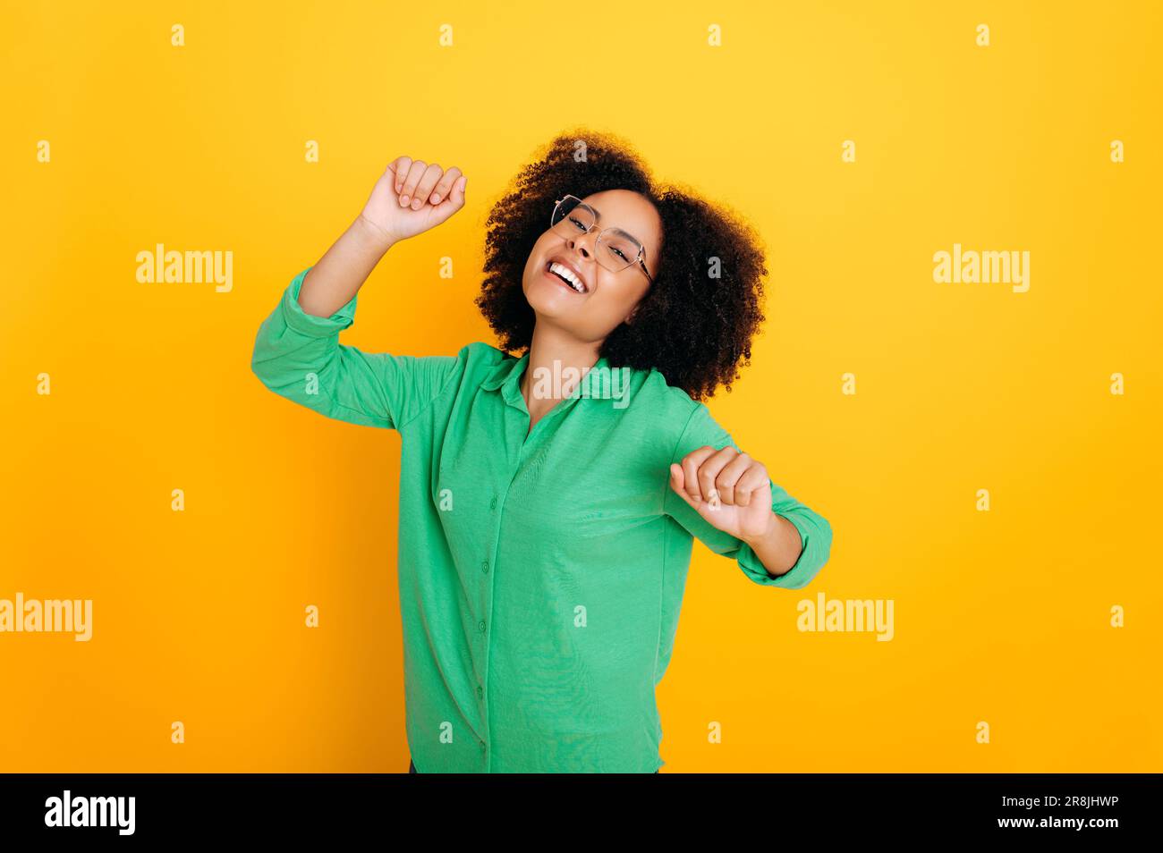 Success, ecstatic. Happy african american or brazilian curly woman with glasses, stand on isolated yellow background, gesturing with fists, laughing, rejoicing, dancing, experiencing joyful emotions Stock Photo