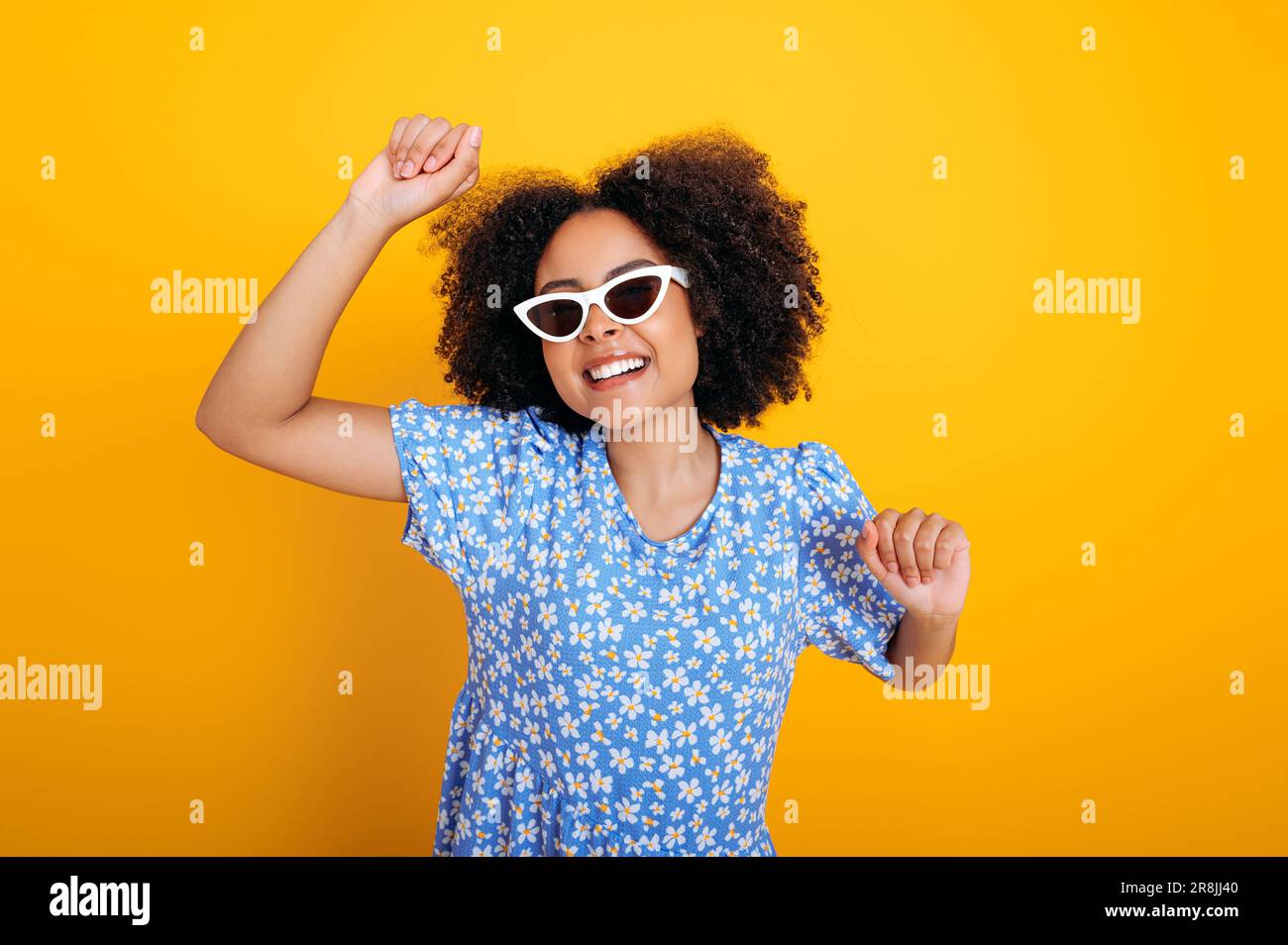Joyful pretty fashionable curly haired brazilian or african american woman, in dress, with sunglasses, listens her favorite music, dancing, relaxing, having fun on isolated orange background, smiling Stock Photo