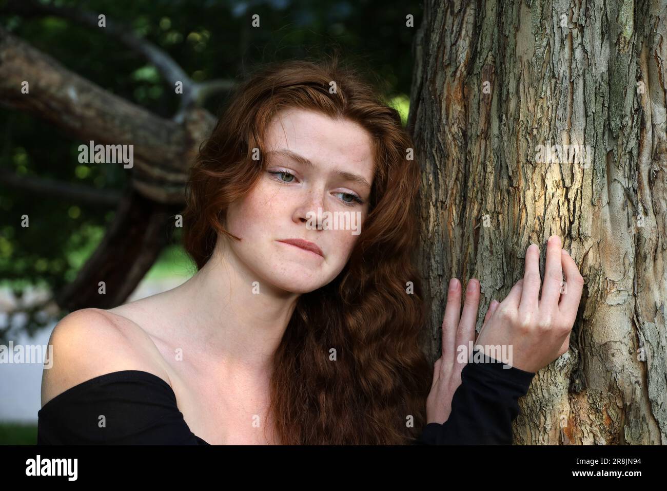 Upset girl with long red hair and freckles standing near the tree in summer park. Depression and sad mood Stock Photo