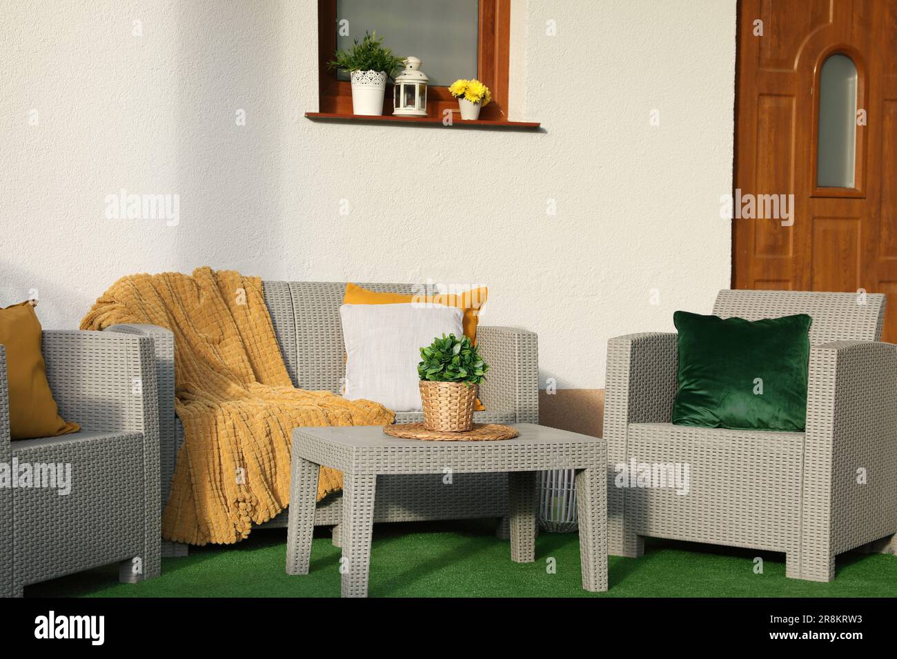 Beautiful rattan garden furniture, soft pillows, blanket and houseplant near white wall Stock Photo