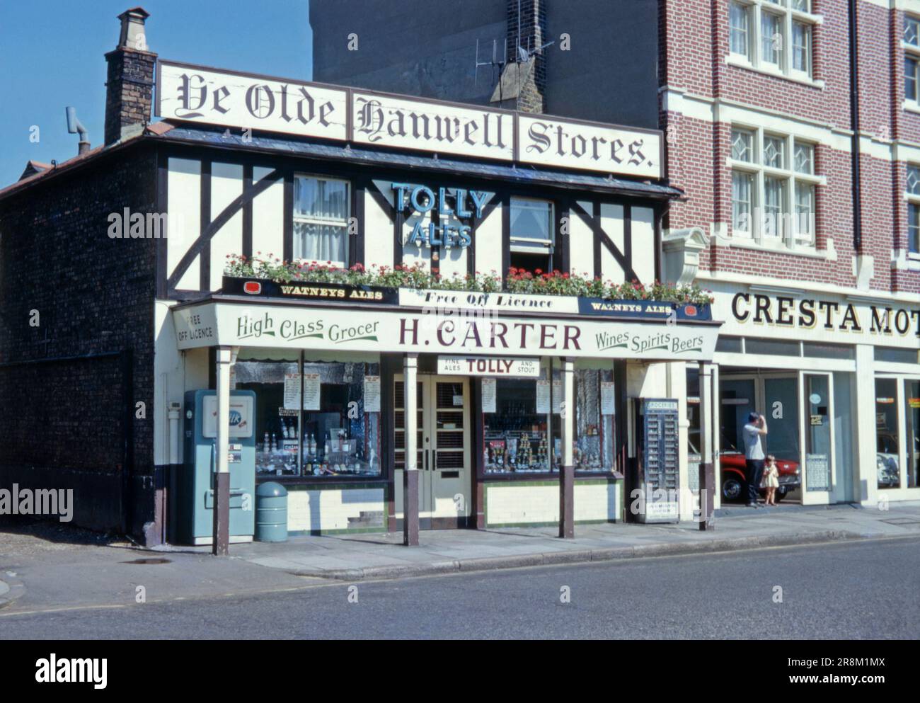‘Ye Olde Hanwell Stores, a ‘high class’ grocer and ‘free off licence’ or general store in the west London suburb of Hanwell, Ealing, England, UK c. 1970. The building sports a mock Tudor look with dark beams on a white façade and has a permanent canopy supported by wooden pillars. As well as groceries, alcohol was sold – the term 'off licence’ means it was not tied to a particular supplier of alcohol. Signs show it sold beers from Watneys and Tolly Cobbold. Vending machines outside allowed grocery items and milk to be sold out of hours – a vintage 1960/70s photograph. Stock Photo